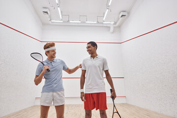happy interracial male friends in sportswear smiling and holding racquets for squash inside of court
