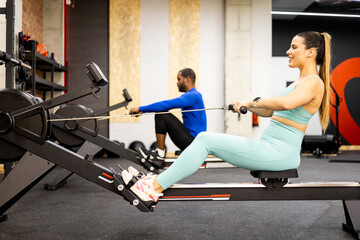 A young couple of athletes of different ethnicities work their bodies with a magnetic air rowing machine. The boys are sitting working their lats with the air rowing machine.