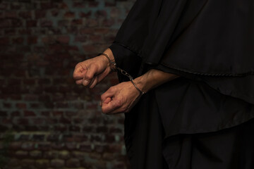 The hands of a monk or priest in handcuffs on the background of a stone wall. Concept: old criminal, criminal liability, religious figure. - obrazy, fototapety, plakaty