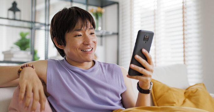 Gen Z queer man non-binary sitting easy relax happy read news on web page app at home sofa. Asia people LGBT young adult guy play web3, store shop e-commerce buyer, virtual retail network cyber paying