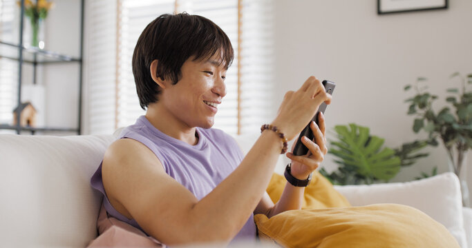 Gen Z queer man non-binary sitting easy relax happy read news on web page app at home sofa. Asia people LGBT young adult guy play web3, store shop e-commerce buyer, virtual retail network cyber paying - Powered by Adobe