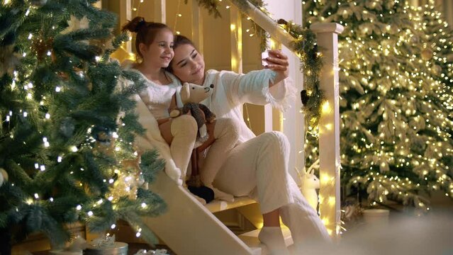 Two girls make a selfie with festive Christmas decoration at the background. Kids wearing white clothes, smile on camera. High quality 4k footage