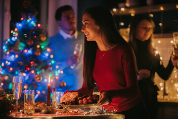 Smiling young woman serving holiday dishes on table while her friends celebrating New Year 