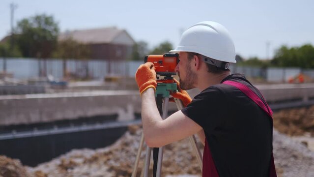 Professional Construction Worker Wearing Hard Hat and Safety Glasses Uses Theodolite at Modern Industrial Manufacturing Facility. Man with optical instrument doing land surveying work.