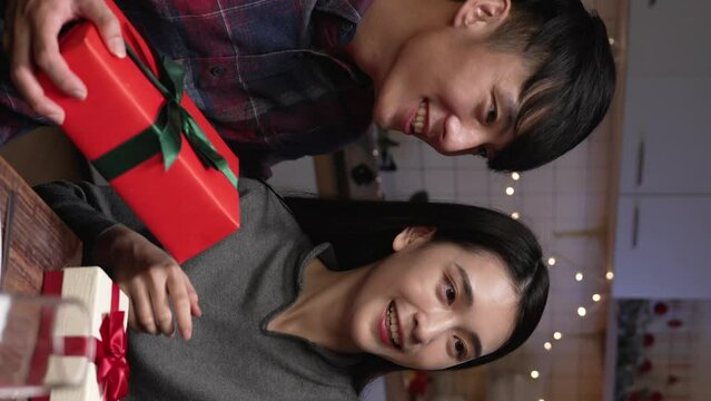 Vertical Screen: Happy asian korean couple exchanging gifts with each other at home in decorated kitchen. cute lovers taking self photo with present box during xmas eve celebration
