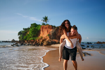 Couple enjoys piggyback ride on tropical beach sunset. Man carries joyful woman on back, strolling along shoreline. Romantic travelers at seaside, exotic holiday escape. Playful duo on sandy coast.
