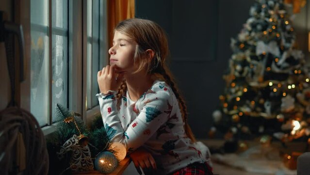 Little girl looking through the window, expecting Santa Claus for Christmas, dreaming and imagining wishes in the living room with a decorated Christmas tree