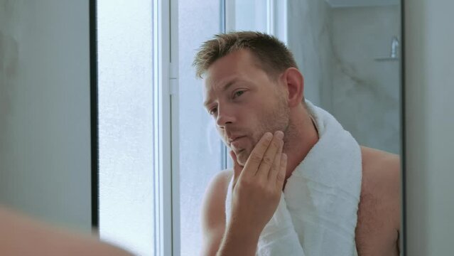 Young handsome young man toching his bristle on the face standing in the bathroom with a towel and looking at mirror. Preparation for daily morning routine male hygienic beauty procedure