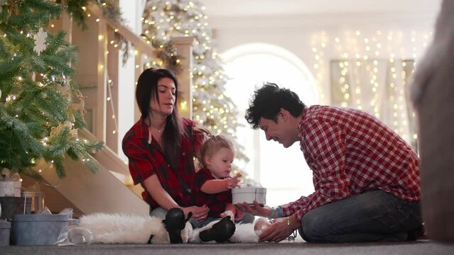 Happy parents give a gift to their baby girl while she ignores it and plays with beads. Young family unwrap Christmas presents in the house full of lightings. High quality 4k footage