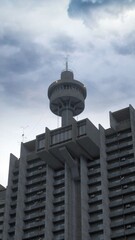 Brutalist architecture. A tall concrete residential estate with a tower on top of it's side and a grey cloudy sky in the background. Photorealistic 3D illustration.