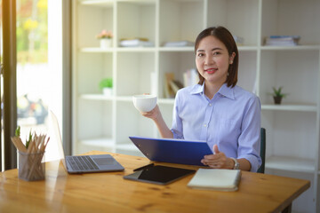 Businesswoman sitting holding a document clipboard while drinking coffee at the table in the office.