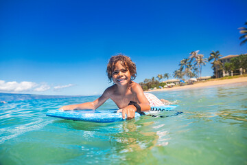Smiling diverse young boy boarding in the beautiful blue ocean. Enjoying a fun day playing at the beach while on vacation in Hawaii