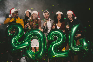 Joyful people in Christmas hats holding 2024 numbers while celebrating New Year in night club