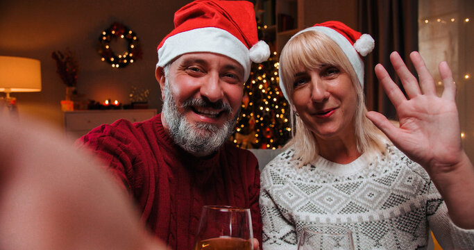 POV of happy senior wife and husband wearing Santa hats sitting at decorated home on Christmas Eve with glasses of wine and speaking on video call in festive mood celebrating New Year. Close up