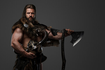 A fierce bearded Viking warrior in fur and light armor, with a helmet attached to his belt, holding...