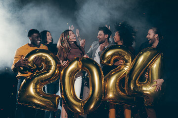Joyful young people holding 2024 gold numbers while celebrating New Year in night club together