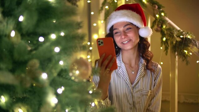 Young caucasian woman in Christmas hat talks on the telephone. Girl wearing pajama set has a video conference. High quality 4k footage
