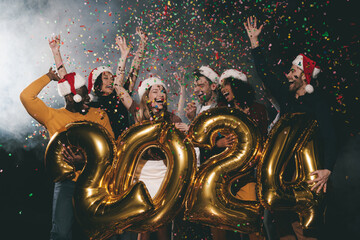 Joyful people in Christmas hats celebrating 2024 New Year while throwing confetti in night club