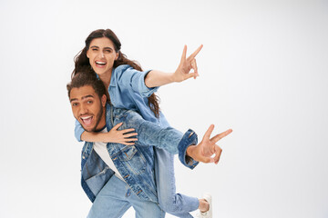 overjoyed couple in trendy denim wear showing victory signs while having fun on grey, piggybacking