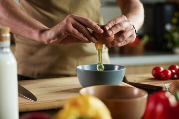 Closeup of unrecognizable man standing by kitchen table cracking egg into bowl