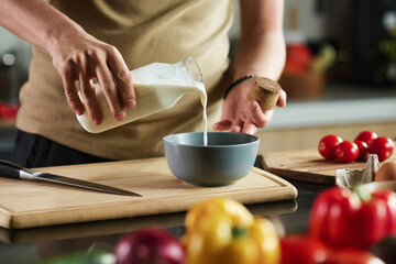 Closeup of unrecognizable man adding milk from glass bottle in bowl while standing by kitchen table