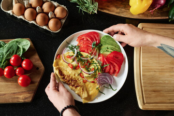 Flat lay shot of plate with omelet and vegetables held by male hands over black kitchen table background with foodstuff and kitchenware around
