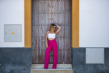 South American woman, young, beautiful and blonde with white top, pink pants and sunglasses, posing in the middle of a wooden door, in sensual and provocative attitude. Trend concept, beauty, fashion