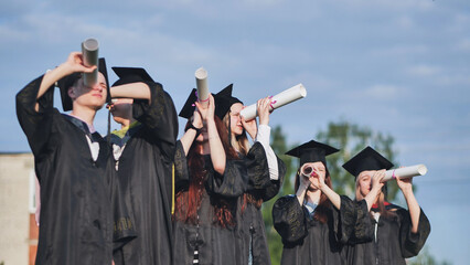 Cheerful graduates on a sunny day look through diplomas like a telescope.