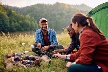Group of happy hikers roasting marshmallows on campfire in front of their tent.