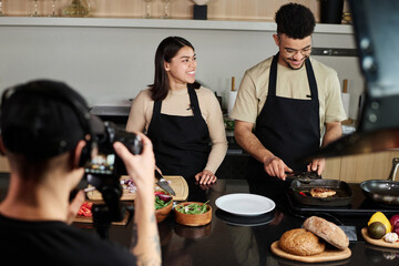 High angle medium shot of young middle eastern man and hispanic woman cooking food and laughing while being filmed by cameraman