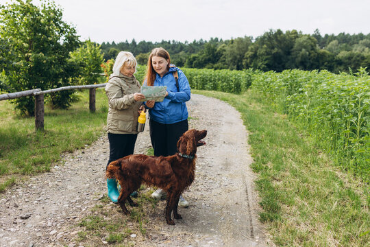 Two happy Womans in active trekking clothes with map having a halt after hiking. Travel and active lifestyle concept, full body. Travel and hiking with pet.