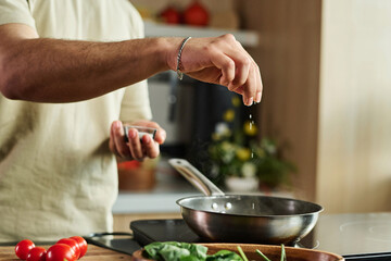 Closeup of hands of unrecognizable man adding salt to meal in pan on kitchen stove