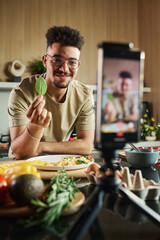 Waist up portrait of smiling young middle eastern blogger at kitchen showing leaf of spinach to...
