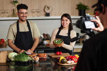 Over shoulder medium shot of young middle eastern man and hispanic woman smiling and showing bowl of arugula to camera while being filmed by cameraman - Powered by Adobe