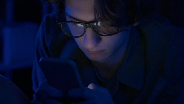 Young man with glasses is texting on his smartphone lying in bed at home at night . Technology, internet, communication and people concept.