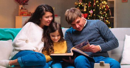 Fototapeta premium Happy Caucasian family with small kid sitting at cozy decorated room in festive mood reading book together on Christmas Eve. Happy holidays. New Year celebration concept. Winter holidays season