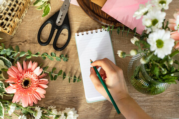 Top view of crop anonymous female florist standing at table surrounded by flowers and taking notes on notebook while working in floral shop