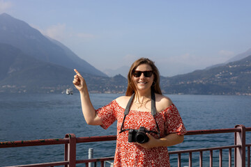 Happy brunette 30s woman enjoying summer holidays. Concept of vacation and travelling. Varenna, Italy. Lake Como. Redhead girl pointing finger and holding a camera outdoors at summer day