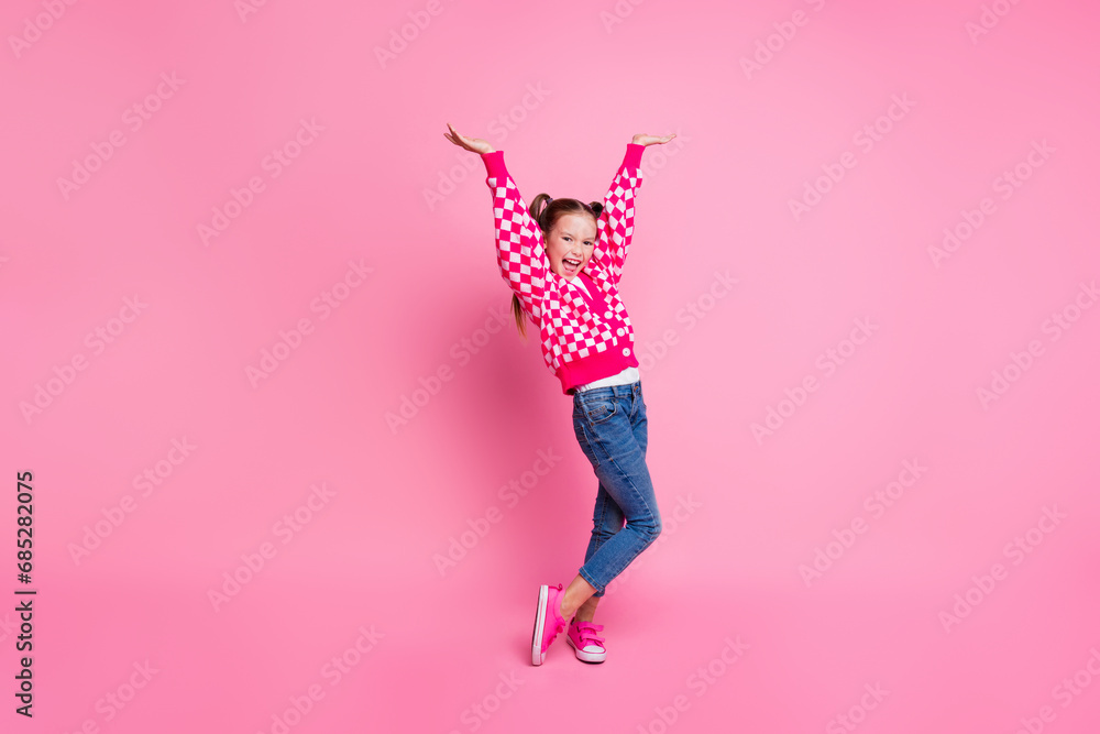 Wall mural Portrait of overjoyed girl with fluttering ponytails wear print sweater denim pants raisng hands up isolated on pink color background