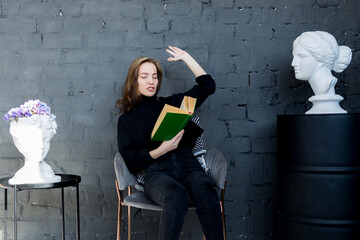 a young woman is sitting on a chair against a gray wall reading a green book waving her arms