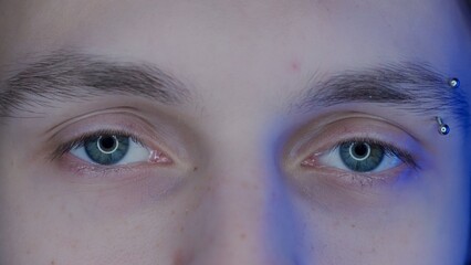 Eyes of young guy with eyebrow piercing macro shot. Blue light from a smartphone, computer screen...