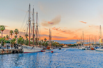 Evening seascape at Port Vell with a pirate galleon and many yachts against the backdrop of an orange-pink-blue sky in Barcelona, Spain. Beautiful cityscape of the Mediterranean coastline at sunset - obrazy, fototapety, plakaty