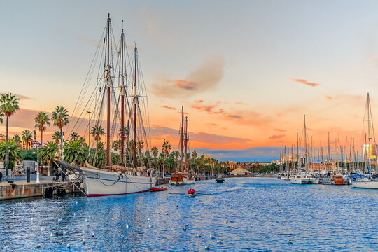 Evening seascape at Port Vell with a pirate galleon and many yachts against the backdrop of an orange-pink-blue sky in Barcelona, Spain. Beautiful cityscape of the Mediterranean coastline at sunset