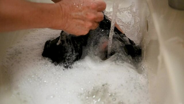 A woman's hands are washing black clothes in the bathroom sink with bar soap.
