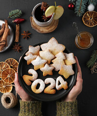 Cookies in the form of numbers 2024 and stars on the dark plate holding by hands and dark...