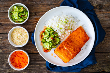 Seared salmon steak with boiled white rice and sliced cucumber on wooden table
