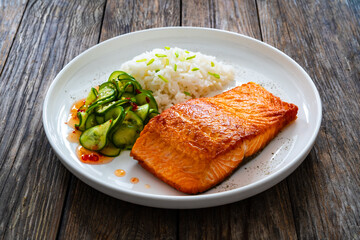 Seared salmon steak with boiled white rice and sliced cucumber on wooden table
