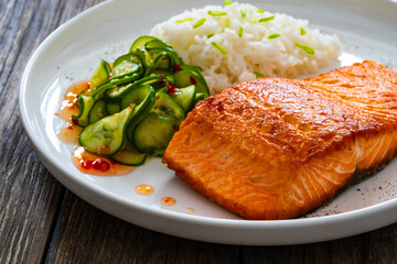 Seared salmon steak with boiled white rice and sliced cucumber on wooden table
