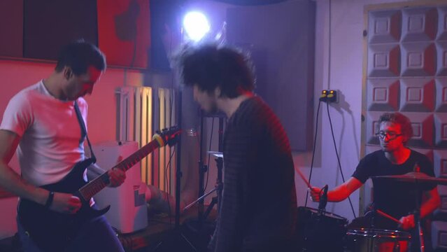 Singer and musicians of a rock band play heavy music in the rehearsal room. The vocalist is headbanging to the beat of the music and singing the song to the accompaniment of a guitarist and a drummer.
