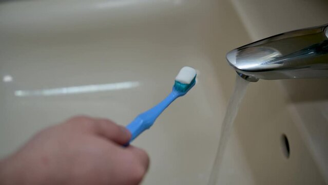 A man's hand opens the tap of the faucet with one hand, while holding a toothbrush with toothpaste on it in the other. He positions it under the running water, rinsing it off.
